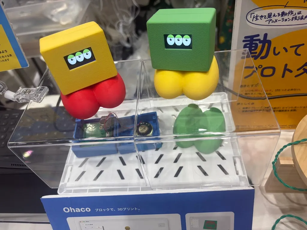 Yellow and green Sankome robots, with their electronics visible under a clear stand and an Ohaco panel in front