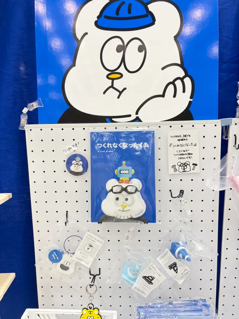 ペグボードに掛けられた絵本『つくれなくなったくま』。背景に大きなしろくまのイラスト、横にしろくまのピンバッジやキーホルダー類